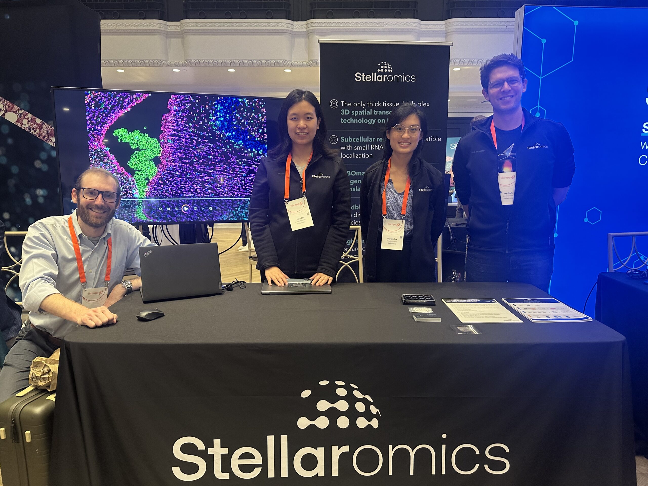 Four employees behind a table with a Stellaromics tablecloth on it, and a large screen displaying images from Pyxa behind the table.