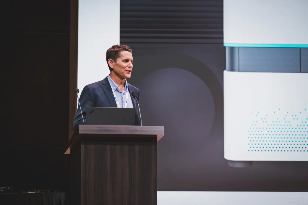 Stellaromics CEO Todd Dickinson standing behind a podium with a laptop. Behind him is a portion of a large screen, with an image of Pyxa.