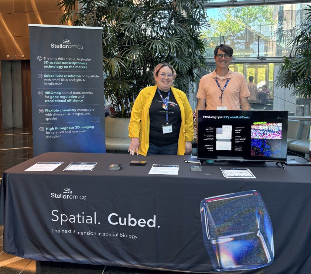 Two smiling employees standing behind a long table, which is covered in a Stellaromics-branded tablecloth, has a monitor displaying images of Pyxa, and with a pop-up banner listing the use cases for Stellaromics behind.